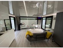 Innenraum eines modernen Hausboots mit viel Glas: Der Raum verfügt über große Fenster und Glastüren mit Blick auf einen See, eine graue Couch mit gelben Kissen, eine minimalistische Küche und einen kleinen Esstisch. Helle Boden- und Wandfarben betonen das lichtdurchflutete Ambiente.