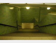 Treppe in einer U-Bahn-Station mit grün gefliesten Wänden, einfachen Metallhandläufen und warmem, gelblichem Licht von Deckenlampen. Am unteren Ende der Treppe ist ein gefliester Gang erkennbar.
