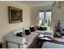 Wohnzimmer mit einer weißen Eckcouch, dekoriert mit verschiedenfarbigen Kissen. Über der Couch hängt ein Gemälde in einem goldenen Rahmen. Auf der rechten Seite befindet sich ein großes Fenster mit Blick auf einen Garten. Ein Kronleuchter hängt von der Decke, und ein Beistelltisch sowie ein Teppich ergänzen die Einrichtung.