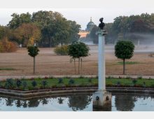 Eine Parklandschaft mit einem rechteckigen Wasserbecken im Vordergrund, eingefasst von Blumenbeeten und kleinen Bäumen. Im Hintergrund steht eine weiße Säule mit einer Büste, dahinter weitläufige Wiesen, Bäume und ein historisches Gebäude mit grüner Kuppel, teilweise in Nebel gehüllt.