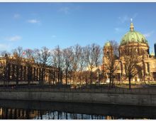 Blick auf den Berliner Dom (rechts) mit seiner grünen Kuppel und das Alte Museum (links) mit einer Säulenfassade, eingerahmt von kahlen Bäumen entlang eines Flussufers unter klarem, blauem Himmel.