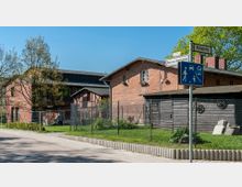 Straßenszene mit roten Backsteingebäuden im Hintergrund, auf einem umzäunten Grundstück mit Gartenfläche und einem Holzschuppen. Im Vordergrund steht ein Verkehrsschild für eine Verkehrsberuhigte Zone, und Straßenschilder weisen auf die „Königsweg“ und „Robert-von-Ostertag-Straße“ hin.