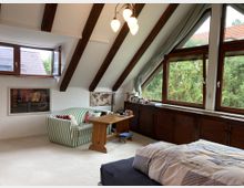Ein Schlafzimmer unter einem Dachgeschoss mit sichtbaren Holzträgern, großen Fenstern mit Ausblick auf grüne Bäume und einer Einrichtung, die ein gestreiftes Sofa, einen Holztisch und ein Bett umfasst. Unter den Fenstern befinden sich Holzschränke, an der Wand hängt ein Gemälde, und der Raum ist mit Teppichboden ausgelegt.