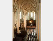 Innenraum einer gotischen Kirche mit spitz zulaufenden Bögen, großen Glasfenstern und weißen Säulen. Die Einrichtung umfasst Holzbänke, eine reich verzierte Kanzel, Kronleuchter und einen Altarbereich im Hintergrund.
