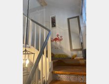 Holztreppe in einem Wohnraum mit weißem Geländer, dekoriert mit Flamingo-Wandbildern, einem großen Wandspiegel und einer Hängelampe. Rechts neben der Treppe befindet sich eine kleine Sitznische mit Kissen und einer Decke.