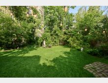 Grüne Gartenfläche mit dichtem Baumbewuchs im Hintergrund, mehreren hängenden Lichterketten und zwei Holzbänken an der Rückseite. Der Garten ist von Schatten durchzogen und enthält eine kleine Statue am Rand sowie einen gepflasterten Weg im Vordergrund.