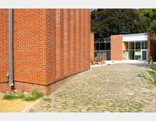 Ziegelbau mit modernen architektonischen Elementen, flankiert von einem gepflasterten Weg und einer Terrasse mit Holztischen und Stühlen. Im Hintergrund befinden sich Glas- und Metallkonstruktionen sowie Bäume.