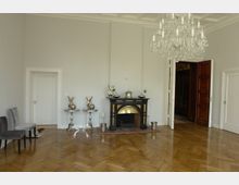 Ein eleganter Raum mit hellem Parkettboden, einem schwarzen Kamin mit goldenen Details in der Mitte sowie dekorativen Metallgefäßen auf kleinen Tischen daneben. Rechts befindet sich eine geöffnete Holztür, links und rechts weiße Türen, und ein großer Kronleuchter hängt von der Decke.