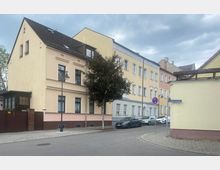 Straßenansicht eines Wohnviertels mit mehrstöckigen, hellfarbigen Gebäuden im Altbaustil, gepflastertem Gehweg und parkenden Autos entlang der Straße. Ein Straßenschild mit der Aufschrift "Grübenstraße" ist an einer Hausecke zu sehen.