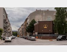 Straßenszene in einer Wohngegend mit parkenden Autos auf beiden Seiten; rechts ein Backsteingebäude mit einem goldenen Symbol einer Krone und Sternen an der Fassade, dahinter weitere mehrstöckige Gebäude mit Balkonen. Im Hintergrund Bäume und bewölkter Himmel.