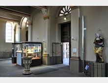 Innenaufnahme einer Kirche mit einem Taufbecken im Vordergrund, einer Vitrine mit Broschüren und Büchern zur rechten Seite und einer Holzstatue auf einem Sockel daneben. Im Hintergrund befinden sich ein offener Eingang mit großen Holztüren und kunstvoll gestaltete Rundbogenfenster.
