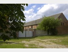 Ehemaliges landwirtschaftliches Gebäude aus roten Ziegelsteinen mit einem grauen Schrägdach, umgeben von einer Steinmauer und grüner Vegetation; im Vordergrund Wiese und ein unbefestigter Weg.