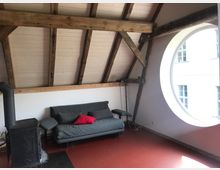 Wohnraum in einem Dachgeschoss mit sichtbaren Holzbalken und einer großen runden Fensteröffnung. Im Raum stehen ein graues Sofa mit roten und grauen Kissen sowie ein schwarzer Holzofen auf einem roten Bodenbelag.