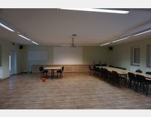 Ein schlicht eingerichteter Seminarraum mit Holzfußboden, rechteckigen Tischen und grünen Stühlen. An der Wand befinden sich zwei weiße Tafeln, eine Projektorhalterung hängt von der Decke, und ein roter Ball liegt auf dem Boden.