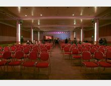 Ein Veranstaltungsraum mit Reihen von roten Stühlen, ausgerichtet auf eine Bühne im Hintergrund. Der Raum hat eine helle Deckenbeleuchtung, Holzwände und eine niedrige Decke mit sichtbaren Stützsäulen.