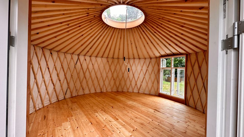 Innenraum einer runden Jurte mit Holzfußboden, hellen Wänden und einem kuppelförmigen Dach mit sichtbaren Holzstreben. Im Dach befindet sich eine runde Öffnung, und an der Seite sind Fenster mit Blick ins Grüne.