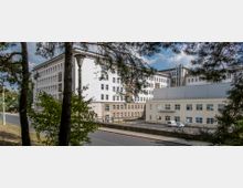 Großes Verwaltungs- oder Schulgebäude in heller Putzfassade mit mehreren Stockwerken und vielen Fenstern, flankiert von Bäumen im Vordergrund. Im Umfeld befinden sich eine Straße, ein Gehweg sowie Parkplätze mit weißen Fahrzeugen.