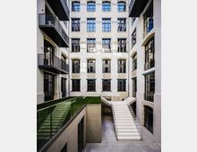 Innenhof eines mehrstöckigen Gebäudekomplexes mit heller Ziegelfassade, großen Sprossenfenstern und seitlichen Balkonen. In der Mitte führen eine breite Treppe und eine Rampe mit Glasgeländern zu einer tieferliegenden Ebene, daneben liegen schmale Rasenflächen.