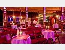 Innenansicht eines nostalgischen Varietézeltes mit Holzboden, runden Tischen und roten Stühlen. Die Tische sind mit weißen Tischdecken, roten Auflagen und Kerzen dekoriert, umgeben von Spiegeln, Säulen und warmem Licht.