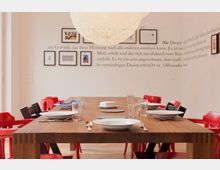 Ein modern gestalteter Essbereich mit einem großen Holztisch, der mit Geschirr, Gläsern und Besteck eingedeckt ist, umgeben von roten und schwarzen Stühlen. An der weißen Wand im Hintergrund befinden sich eingerahmte Bilder und ein Wandtext.