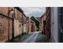 Schmale Pflasterstraße in einer historischen Altstadt, gesäumt von Backstein- und Fachwerkhäusern sowie Laternen. Im Hintergrund sind bewachsene Fassaden und Dächer zu sehen, unter einem teils bewölkten Himmel.