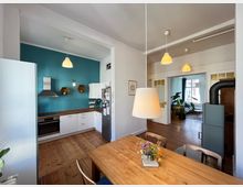 Wohnküche mit Holzboden und einer offenen Verbindung zum Wohnbereich. Die Küche umfasst weiße Schränke, eine blaue Wand, eine Dunstabzugshaube und Hängelampen; im Vordergrund steht ein Holztisch mit einer Blumenvase, im Hintergrund befindet sich ein Kaminofen und große Fenster mit Pflanzen.