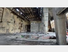 Industriehalle im verlassenen Zustand mit offenliegendem Betonboden, Backsteinwänden und sichtbaren Stahlträgern an der Decke. Große Fenster und beschädigte Bereiche im Boden sowie rostige Metallelemente prägen das Erscheinungsbild.