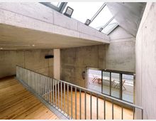 Moderne Innenraumansicht mit Sichtbetonwänden, einer hölzernen Bodenfläche und einer Metallbrüstung. Das Obergeschoss führt zu einem Bereich mit großen Glas-Schiebetüren, die auf eine Terrasse mit Holzmöbeln hinausgehen.