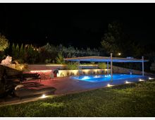 Beleuchteter Außenbereich bei Nacht mit einem Swimmingpool unter einer modernen Überdachung. Im Vordergrund stehen Liegestühle und Sitzmöbel auf einer Terrasse, umgeben von dezent beleuchteten Pflanzen und einer niedrigen Mauer.