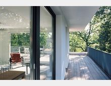 Moderne Terrasse mit Holzboden, angrenzend an ein Wohngebäude mit Glas-Schiebetüren. Der Blick zeigt angrenzende Bäume und eine teils möblierte Sitzecke im Innenbereich.