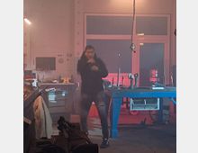 Ein Werkstattraum mit einer Person, die sich in der Mitte bewegt. Im Hintergrund sind Arbeitsbänke, Werkzeuge, ein Schraubstock und eine Tür mit Glaseinsatz, beleuchtet von rötlichem Licht, zu sehen.