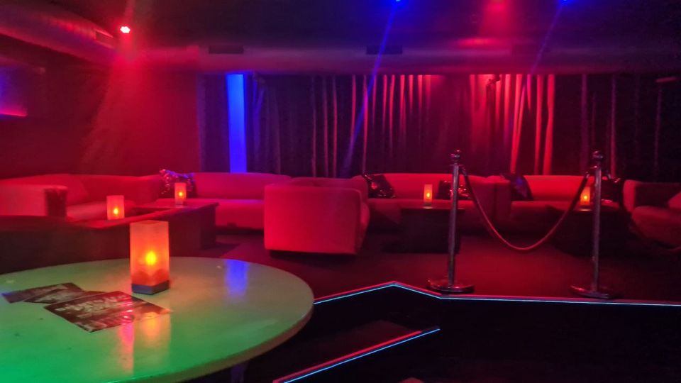 Gedimmter Lounge- oder Clubinnenraum mit roten und blauen Deckenlichtern, runden Tischen mit Kerzenlampen und umlaufenden Polstersofas. Vor einer erhöhten Sitzfläche mit Seilabsperrung hängen dunkle Vorhänge an der Rückwand.
