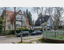 Eine ruhige Wohnstraße mit Kopfsteinpflaster, gesäumt von Einfamilienhäusern mit Satteldächern und hellen Fassaden. Im Vordergrund stehen geparkte Autos und eine grüne Holzbakelemente, während Straßenschilder die Kreuzung „Rohlfstraße“ und „Drygalskistraße“ markieren.