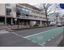 Straßenszene vor dem Einkaufszentrum Boulevard Berlin mit großen Fenstern und mehreren Geschäften im Erdgeschoss. Im Vordergrund sind eine grüne Fahrradspur, Autos, ein Baumbestand und Gehwege mit Fußgängern zu sehen.