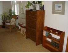 Ein gemütlicher Innenraum mit Holzmöbeln, darunter ein schmaler Bücherschrank mit Büchern und Dekorationen, ein hoher Schrank und ein kleiner Stuhl aus Rattan neben großen Zimmerpflanzen. An der Wand hängt ein gerahmtes Bild, und ein dekoratives Modell eines Segelbootes steht vor dem Fenster.