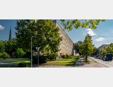 Straßenszene in einer Wohngegend mit einem mehrstöckigen Wohngebäude auf der linken Seite, umgeben von Bäumen und gepflegtem Grün. Im Hintergrund ist ein Kirchturm erkennbar, und auf der rechten Seite führt eine Straße mit geparkten und fahrenden Autos entlang.