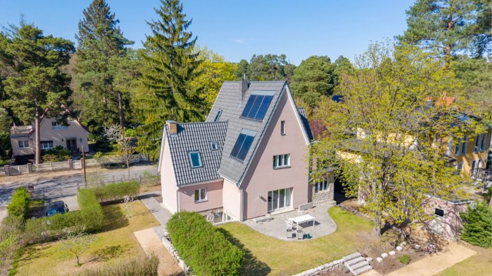 Luftaufnahme eines freistehenden Einfamilienhauses mit steilem grauem Ziegeldach und Dachfenstern, umgeben von Rasenflächen, Hecken und einer gepflasterten Terrasse. Das Haus liegt in einer grünen Wohngegend mit hohen Bäumen und benachbarten Häusern entlang einer Straße.