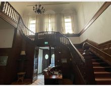 Holzvertäfeltes Treppenhaus in einem historischen Gebäude mit geschwungener doppelter Holztreppe, großen Fenstern mit weißen Vorhängen und einer dunklen Kronleuchter-Decke. Im Erdgeschoss befindet sich eine Eingangstür mit Glaseinsätzen.