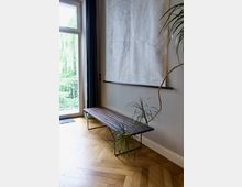 Minimalistisch eingerichteter Innenraum mit hellem Holzboden in Fischgrätmuster, einer schlichten Holzbank mit Metallbeinen, einer Zimmerpflanze und einer großen weißen Karte oder Zeichnung an der Wand. Eine Glastür mit Blick auf einen grünen Außenbereich ist im Hintergrund sichtbar.