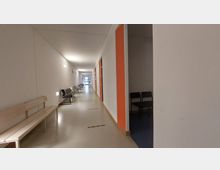 Langer, heller Flur in einem Verwaltungs- oder Klinikgebäude mit weißen Wänden und einem Boden in Beige. Entlang der Wände stehen Bänke und Metallstühle, teilweise mit schwarzem Polster, und Türen mit orangefarbenen Türrahmen führen zu angrenzenden Räumen. Am Ende des Flurs sind eine gläserne Tür und Tageslicht sichtbar.