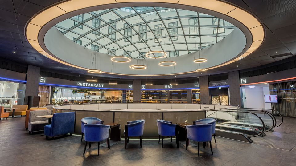 Moderne Innenansicht eines Gebäudes mit einem kreisförmigen Glasdach, das Tageslicht einlässt. Der Raum verfügt über Sitzbereiche mit blauen Sesseln, eine Rolltreppe und Beschilderungen für ein Restaurant und eine Bar.