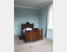 Ein einfaches Schlafzimmer mit hellblauen Wänden, einem hölzernen Einzelbett im antiken Stil, weißem Bettzeug und einem kleinen Nachttisch mit einer Lampe. Der Boden besteht aus hellen Holzdielen, und durch ein Fenster mit weißen Vorhängen fällt Tageslicht in den Raum.