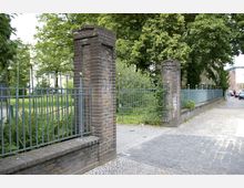 Eingangstor mit Metallzaun und Ziegelpfosten, umgeben von grüner Vegetation auf der linken Seite. Rechts führt ein gepflasterter Bürgersteig an der Zaunanlage vorbei, im Hintergrund sind Gebäude und parkende Autos sichtbar.