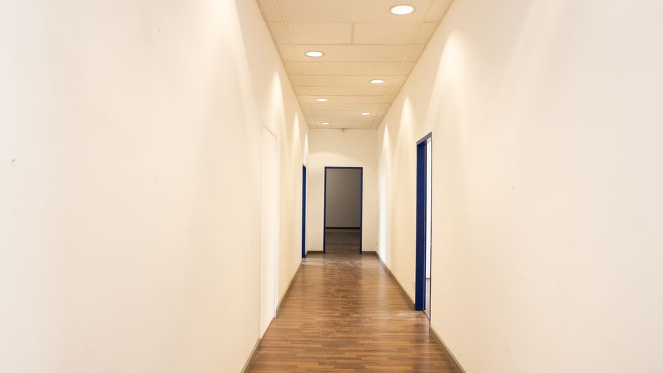 Langer, schmaler Innenflur mit weißen Wänden, Holzfußboden und abgehängter Decke mit runden Einbauleuchten. Entlang des Gangs befinden sich mehrere Türen mit dunkelblauen Rahmen, am Ende ist ein offener Durchgang zu einem weiteren Raum sichtbar.