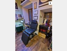 Innenraum eines Tattoo- oder Piercingstudios mit schwarzem Behandlungsstuhl, grünen Rollcontainern und Arbeitsfläche unter einer Stehlampe. An den Wänden hängen gerahmte Zertifikate, daneben ein kleines Fenster mit Jalousien und ein elektrischer Kamin auf einem Holzpodest.