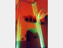Innenraum einer Kirche oder eines historischen Gebäudes mit hohen, farbig angestrahlten Wänden in Orange und Grün. Zu sehen sind Rundbögen, dicke Säulen und romanische Architekturelemente.