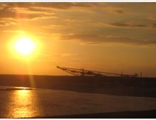 Sonnenuntergang über einer Industrielandschaft mit einem großen Bagger oder Förderband im Hintergrund. Im Vordergrund ist ein Gewässer zu sehen, dessen Oberfläche das Sonnenlicht reflektiert.