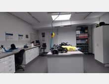 Raum mit technischem Equipment, vermutlich für Atemschutzgeräte-Wartung. Zu sehen sind ein großer Tisch mit Atemschutzgeräten, Schränke, Regale mit Flaschen und Ordnern sowie Arbeitsplätze mit Stühlen und Computern, in einem funktionalen, neutralen Umfeld.