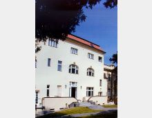 Helles, dreistöckiges Gebäude im klassischen Stil mit einem roten Satteldach und großen, symmetrisch angeordneten Fenstern, darunter Rundbogenfenster im ersten Stock. Eine breite Treppe führt zum Eingang, und der Vordergrund zeigt eine kleine Grünfläche mit Gehwegen.