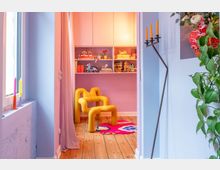 Cozy, colorful reading nook with a bright yellow sculptural lounge chair on a wooden floor, set in front of built-in shelves filled with books and small figurines. A window with sheer curtains, candle holders, and a tall minimalist floor candelabra frame the space, with a pink wall and patterned rug adding color.